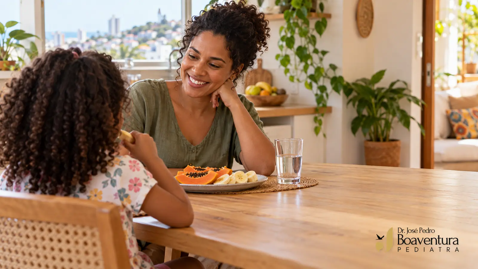 Mãe e criança vista de costas compartilham frutas à mesa, com um copo de água e luz natural.